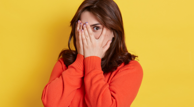 lady in orange top on yellow background peeping out from behind her hands