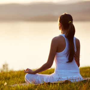 Woman with long dark hair sat on grass overlooking water, sat in yoga positon in white gym pants and vest