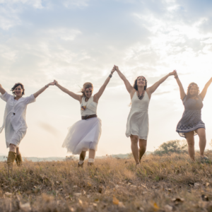 4 women in summery clothes with arms in the air holding hands walking in long grass under a bright cloudy sky