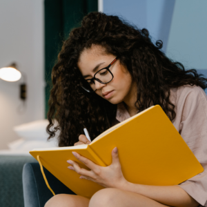 woman in glasses sat with a large yellow notebook in her lap writing