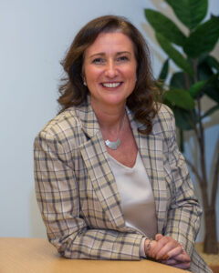 dark haired lady sat at a desk smiling in a checked jacket