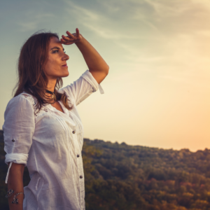 Woman with long hair wearing white shirt with her hand raised to her forehead as she looks ahead