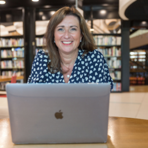 Woman sat in library at her laptop smiling at the camera