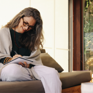 Lady with long hair and glasses sitting with one leg uner her writing in a journal with her back to the glass door