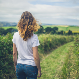 rear view of lady with long hair in white t shirt and jeans walking in the countryside along a grassed path