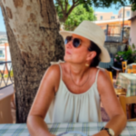 Sharon sitting in reflective mood in a restaurant in the Mediterranean wearing a sun hat and sunglasss